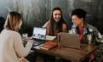 Drei Frauen sitzen am Tisch mit Laptops und lachen