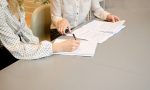 2 Frauen mit hellen Blusen sitzen an grauem Tisch und lehnen sich über Papiere