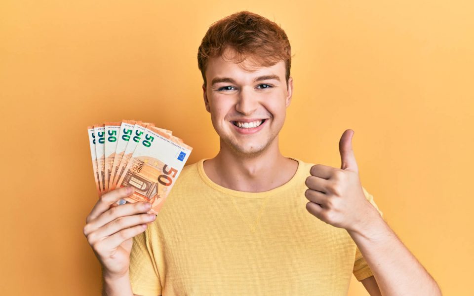 Young caucasian man holding bunch of 50 euro banknotes smiling happy and positive, thumb up doing excellent and approval sign